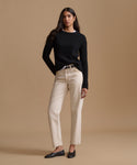 A woman modeling a black ribbed knit sweater and straight-leg beige jeans with black flats, standing against a solid warm beige studio background.