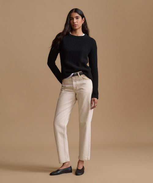 A woman modeling a black ribbed knit sweater and straight-leg beige jeans with black flats, standing against a solid warm beige studio background.