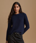 A woman modeling a dark navy ribbed knit sweater with long sleeves and a crew neck, paired with dark jeans, against a solid warm beige studio background.