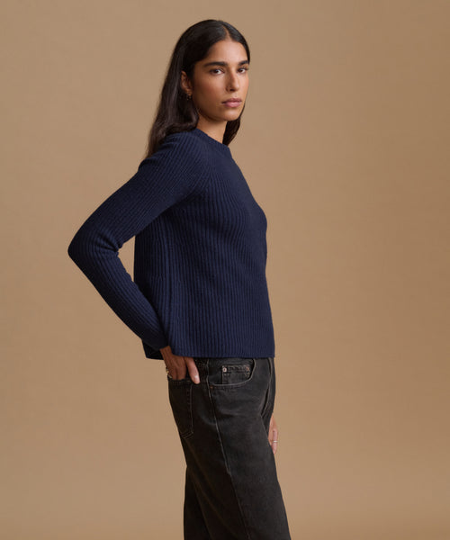 A woman modeling a navy blue ribbed knit sweater with dark charcoal jeans, standing against a solid warm beige studio background.
