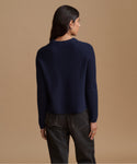 A woman shown from the back wearing a navy blue ribbed knit sweater and dark gray jeans, standing against a solid tan studio background.