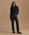 A woman stands against a plain beige studio background wearing a dark navy ribbed knit sweater, dark charcoal gray straight-leg jeans with a black belt, and black slip-on shoes.