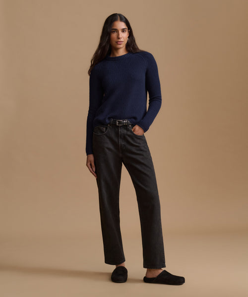A woman stands against a plain beige studio background wearing a dark navy ribbed knit sweater, dark charcoal gray straight-leg jeans with a black belt, and black slip-on shoes.