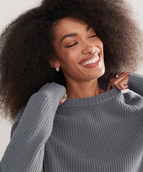 A joyful woman with curly hair and small hoop earrings smiles with her eyes closed, pulling at the collar of a gray Cashmere Fisherman Sweater.