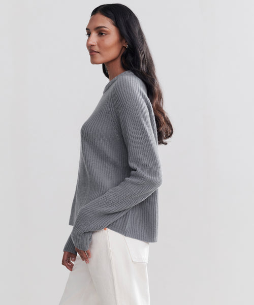 A woman with long dark hair wears the Cashmere Fisherman Sweater in gray and white pants, standing in profile against a plain light background.