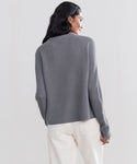 A woman with long dark hair, seen from behind, wears a Cashmere Fisherman Sweater in dusty blue and off-white pants, standing against a plain light background.
