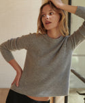 A woman wearing a ribbed knit crewneck sweater in a warm gray color, posing indoors with a neutral background.