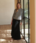 A woman stands barefoot beside a glass door, wearing a light gray knit sweater and a long black pleated skirt.
