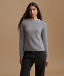 A woman modeling a light gray ribbed knit sweater paired with dark charcoal jeans, against a solid warm beige studio background.