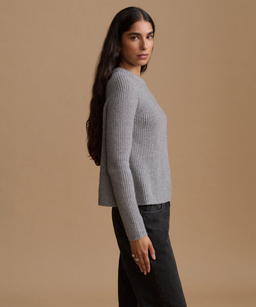 A woman shown in profile wearing a light gray ribbed knit sweater and dark jeans, standing against a solid warm beige studio background.