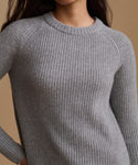 A close-up of a person wearing a light gray ribbed knit crewneck sweater against a warm beige background.