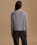 A woman shown from the back wearing a gray ribbed knit sweater and dark jeans, standing against a solid tan studio background.