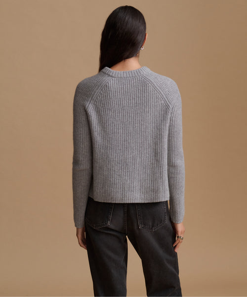 A woman shown from the back wearing a gray ribbed knit sweater and dark jeans, standing against a solid tan studio background.