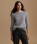 A woman modeling a light gray ribbed knit crewneck sweater, paired with black jeans and a black belt, against a solid beige studio background.