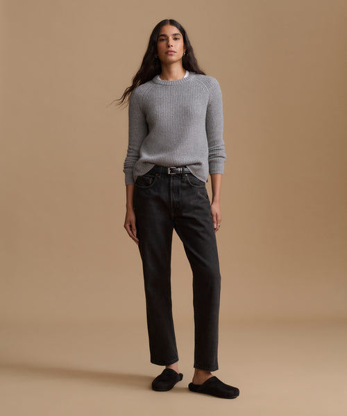 A woman stands against a plain beige studio background wearing a light gray ribbed knit sweater, black straight-leg jeans, and black slip-on shoes.