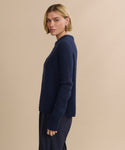A woman with short blonde hair models the Cashmere Fisherman Sweater in navy with matching pants, standing sideways and looking at the camera against a plain beige background.