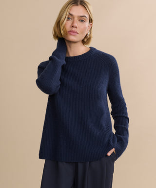 A woman with short blonde hair models the Cashmere Fisherman Sweater in navy and dark pants against a beige background, one hand at her neck and the other in her pocket.