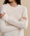 A woman stands with arms crossed, wearing a cozy Cashmere Fisherman Sweater in cream. Gold rings accent her fingers, her face partly out of frame. The background is a soft beige.