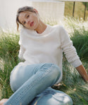 A woman sitting in tall green grass wearing a cream ribbed knit sweater and light blue jeans.