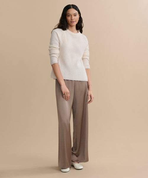 A woman with long, wavy dark hair stands against a beige background wearing an ivory Cashmere Fisherman Sweater, loose taupe trousers, and white shoes. She has a neutral expression and her arms are relaxed by her sides.