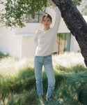 A woman stands outdoors in a grassy area next to a tree trunk, wearing a cream ribbed knit long-sleeve sweater and light blue jeans.
