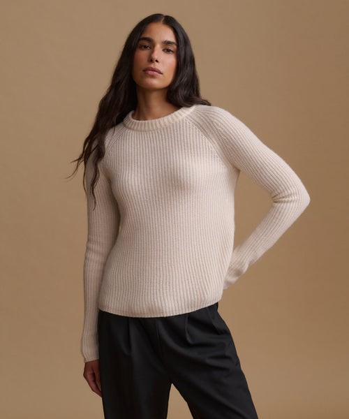 A woman modeling a cream ribbed knit crewneck sweater with black trousers, posed against a solid warm beige studio background.
