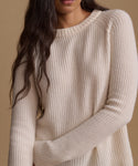 A close-up of a woman wearing a cream-colored ribbed knit sweater against a warm beige background.