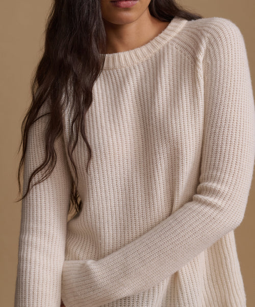 A close-up of a woman wearing a cream-colored ribbed knit sweater against a warm beige background.