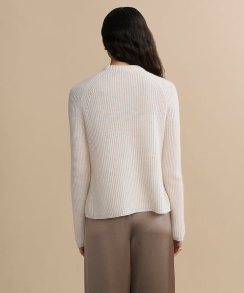 A person with long, wavy dark hair is shown from behind, wearing a Cashmere Fisherman Sweater in ivory and taupe wide-leg pants against a plain beige background.