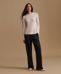 A woman modeling a cream ribbed knit long-sleeve sweater with black wide-leg trousers, standing against a solid beige studio background.