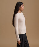 A woman in side profile wearing a cream ribbed knit long-sleeve sweater and black trousers, standing against a solid tan studio background.