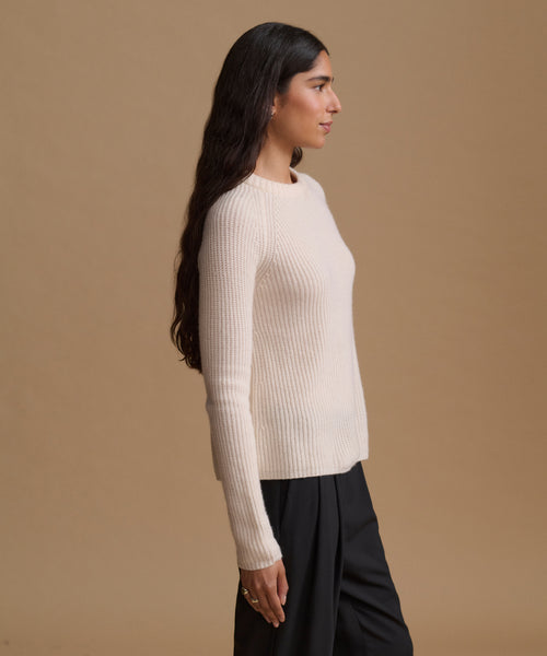 A woman in side profile wearing a cream ribbed knit long-sleeve sweater and black trousers, standing against a solid tan studio background.