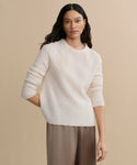 A woman with long, wavy dark hair wears a Cashmere Fisherman Sweater in ivory and loose taupe pants, standing against a plain beige background with a relaxed expression.