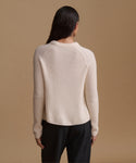 A woman shown from the back wearing a ribbed knit sweater in a light cream color and black trousers, set against a solid warm beige background.