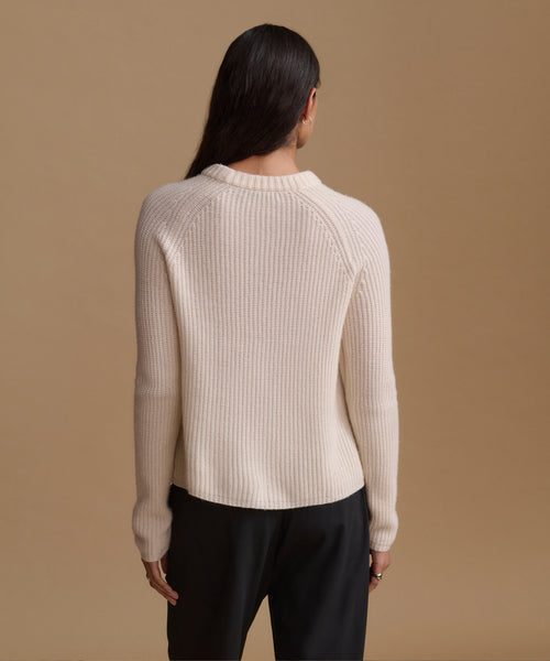 A woman shown from the back wearing a ribbed knit sweater in a light cream color and black trousers, set against a solid warm beige background.