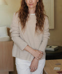 A woman wearing a beige ribbed knit sweater and white pants, standing in a softly lit indoor room with neutral decor.