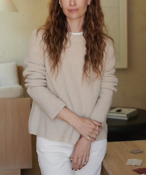 A woman wearing a beige ribbed knit sweater and white pants, standing in a softly lit indoor room with neutral decor.