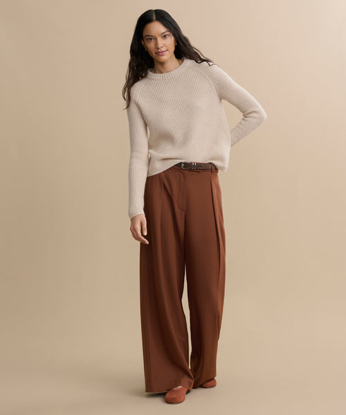 A person with long dark hair wears a Cashmere Fisherman Sweater, wide-leg brown trousers, and brown shoes, standing confidently against a plain beige background.