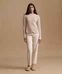 A woman stands against a warm beige studio background wearing a ribbed knit long-sleeve sweater, straight-leg trousers, and slip-on shoes.