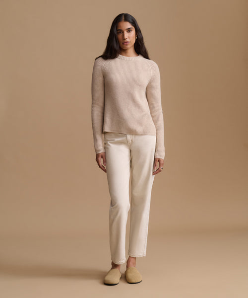 A woman stands against a warm beige studio background wearing a ribbed knit long-sleeve sweater, straight-leg trousers, and slip-on shoes.