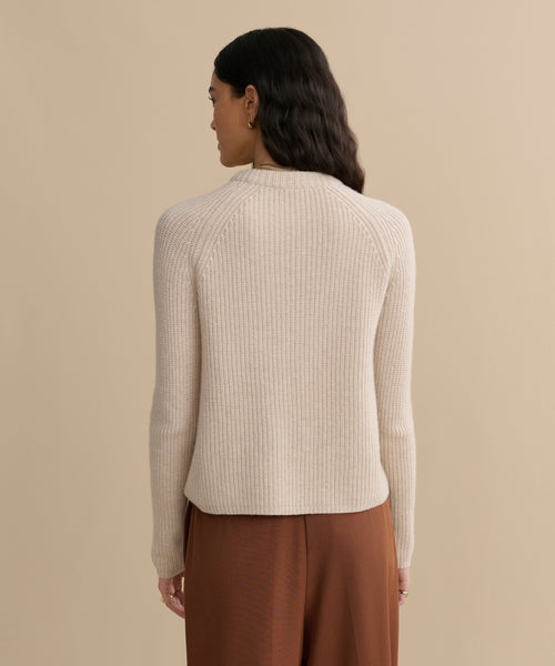 A woman with long, wavy dark hair stands facing away, wearing the Cashmere Fisherman Sweater in ribbed oatmeal and loose brown pants against a plain beige background.