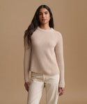 A woman modeling a ribbed knit crewneck sweater in a light beige color, paired with cream-colored pants, against a solid warm beige studio background.