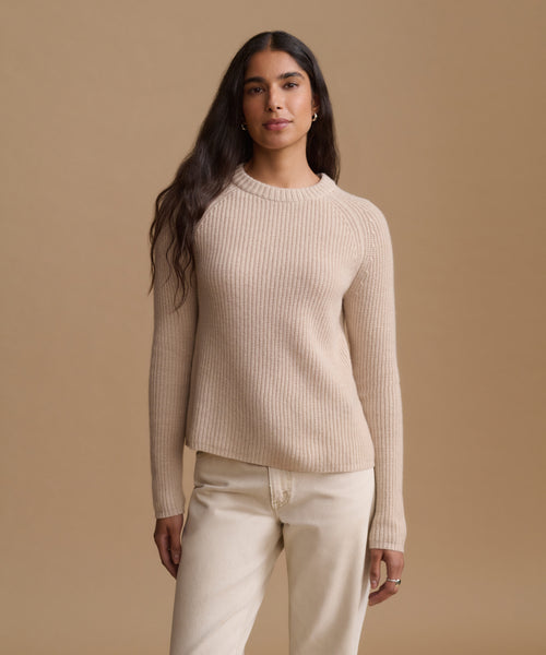 A woman modeling a ribbed knit crewneck sweater in a light beige color, paired with cream-colored pants, against a solid warm beige studio background.
