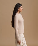 A woman in side profile wearing a cream ribbed knit sweater and matching cream trousers, set against a solid warm beige studio background.