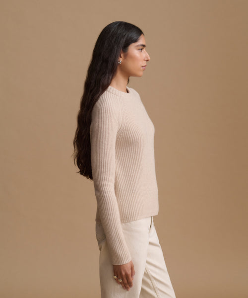 A woman in side profile wearing a cream ribbed knit sweater and matching cream trousers, set against a solid warm beige studio background.