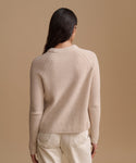A woman shown from the back wearing a ribbed knit sweater in a light beige color, paired with cream-colored trousers, against a solid beige studio background.