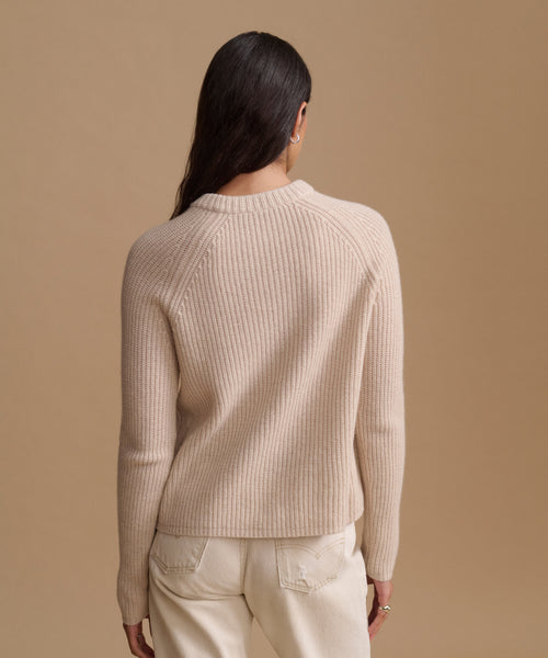A woman shown from the back wearing a ribbed knit sweater in a light beige color, paired with cream-colored trousers, against a solid beige studio background.