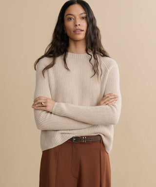 A woman with long wavy hair wears the Cashmere Fisherman Sweater in oatmeal and brown high-waisted belted pants, standing against a plain beige background with her arms crossed.