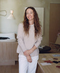 A woman stands in a room wearing a ribbed light beige sweater and white pants. 
