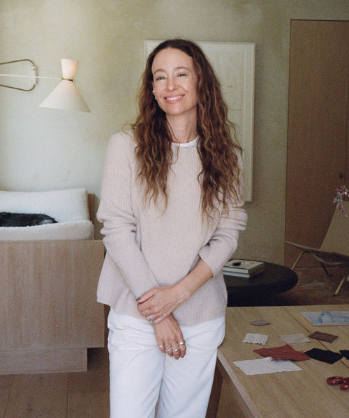 A woman stands in a room wearing a ribbed light beige sweater and white pants. 